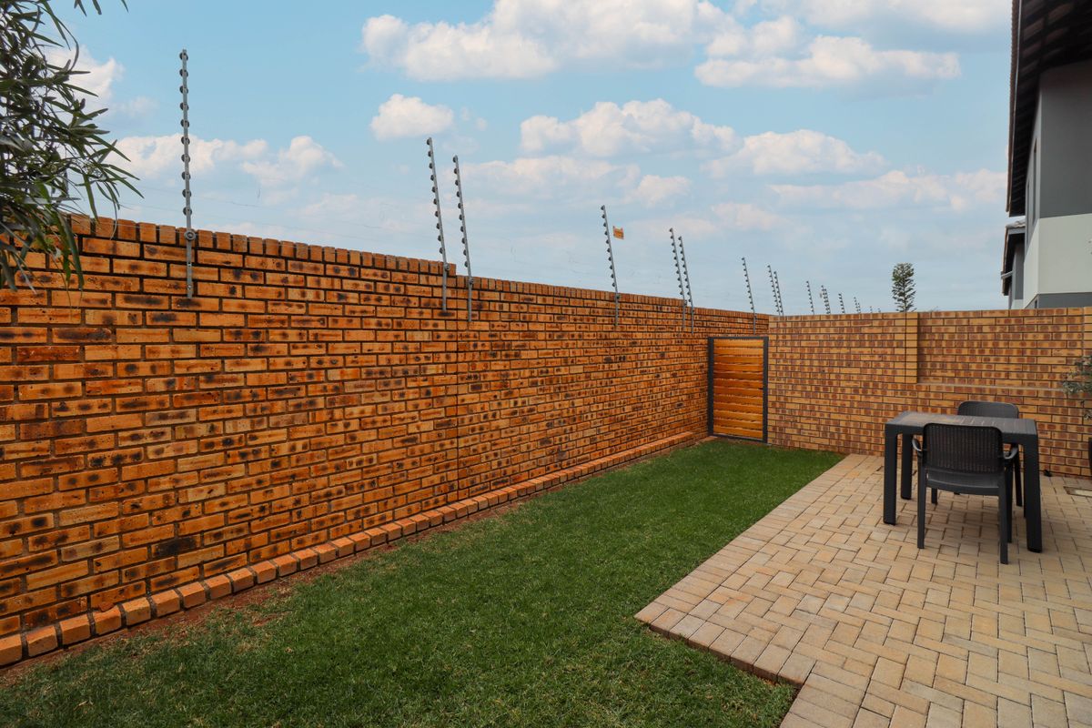 A private rear garden featuring a paved patio area, well-maintained lawn, and a secure face brick boundary wall topped with electric fencing.