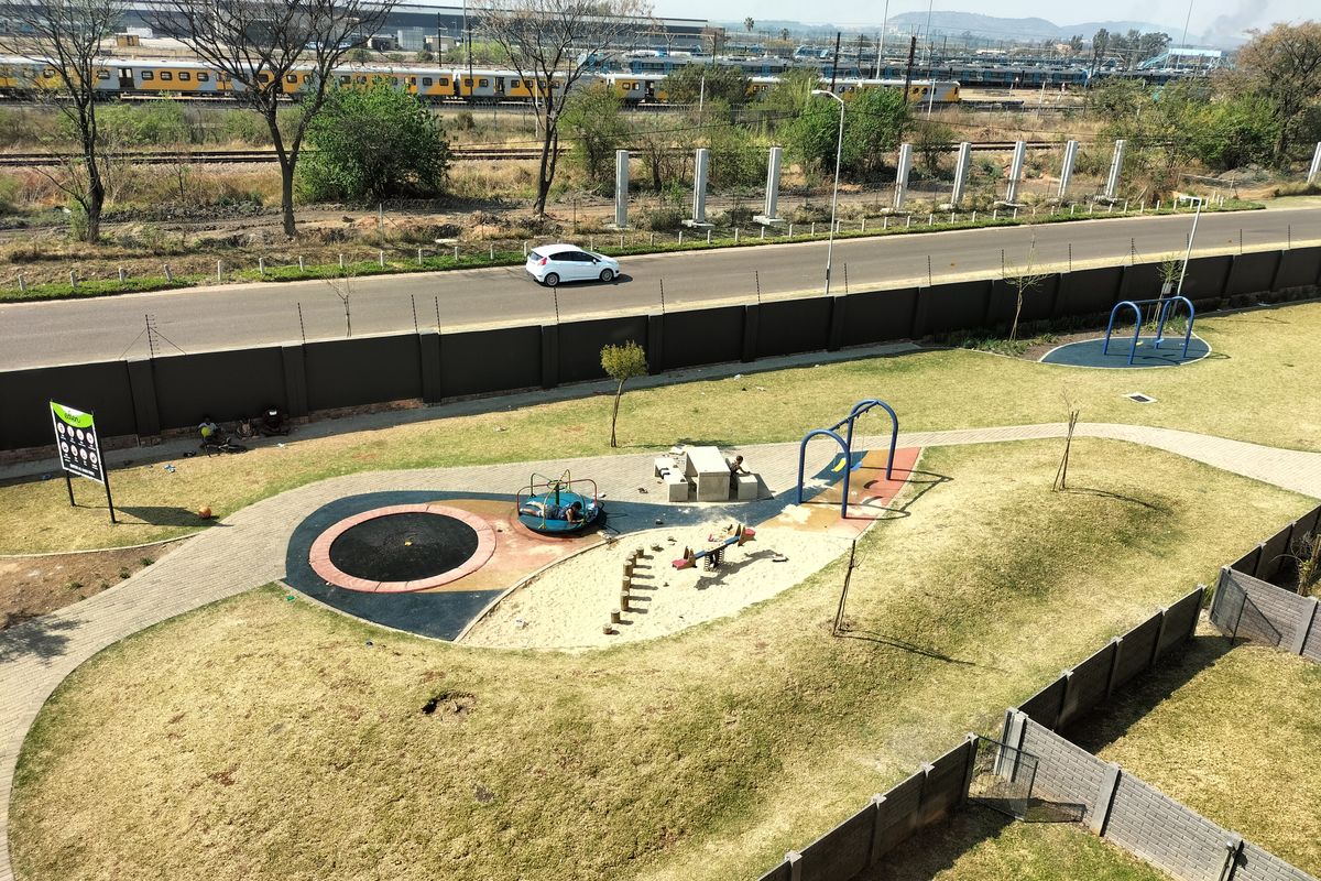 Aerial view of a communal garden area featuring paved walkways, landscaped lawns, and perimeter security fencing with an adjacent road and railway line.