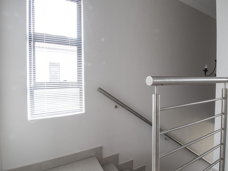 A bright staircase landing featuring a modern stainless steel balustrade and handrail, complemented by tiled steps and a window with venetian blinds.