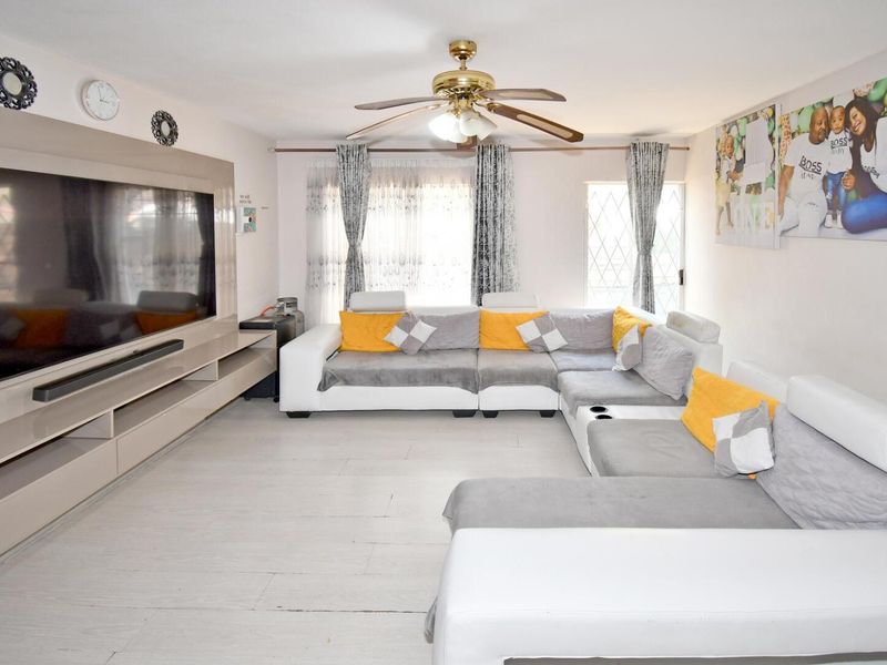 A spacious living room featuring a built-in media unit, ceiling fan, and light-toned laminate flooring with large windows for natural light.