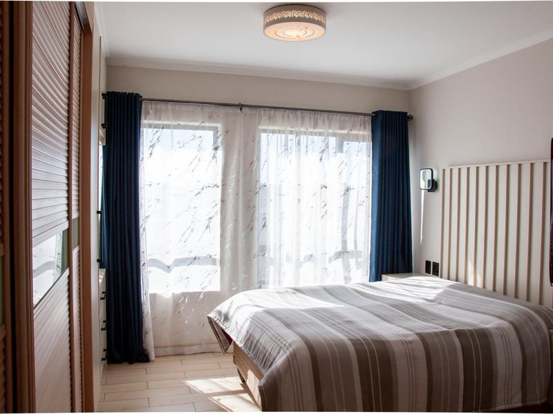 A well-maintained bedroom featuring a built-in wardrobe, slatted headboard, and tiled flooring, illuminated by natural light through large windows.