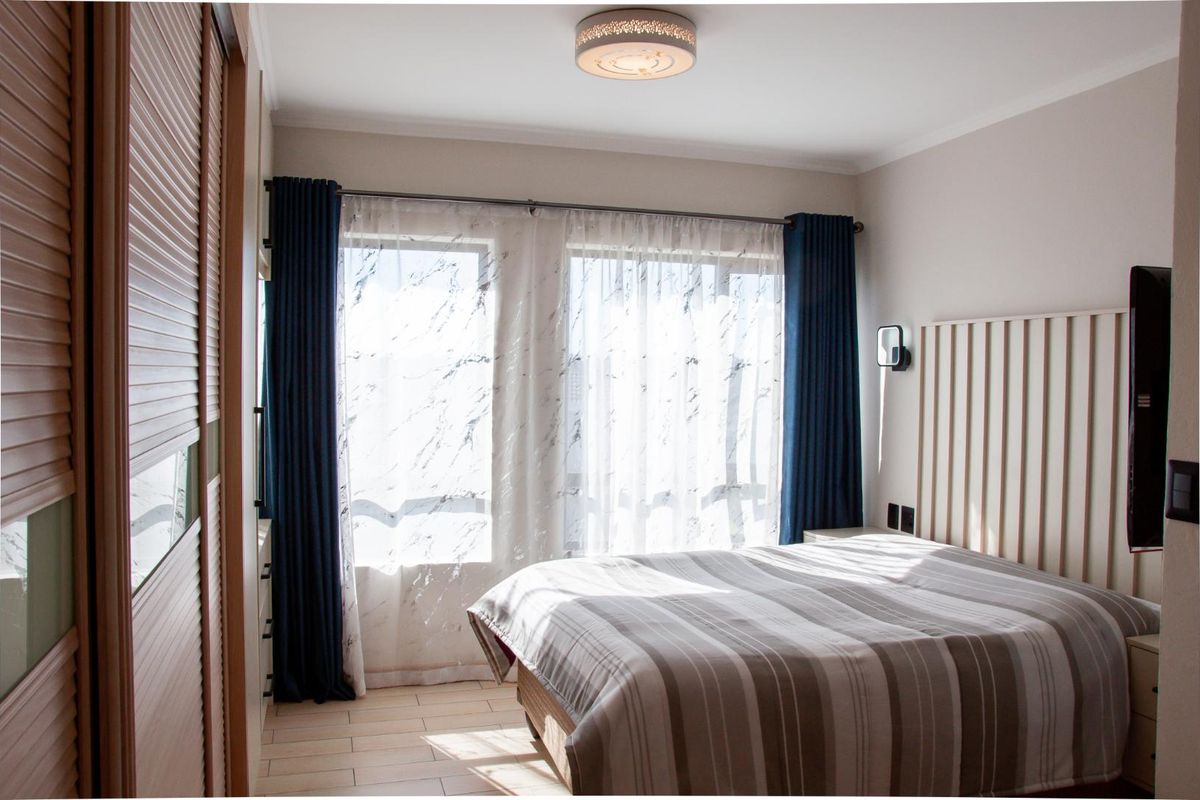 A well-maintained bedroom featuring a built-in wardrobe, slatted headboard, and tiled flooring, illuminated by natural light through large windows.