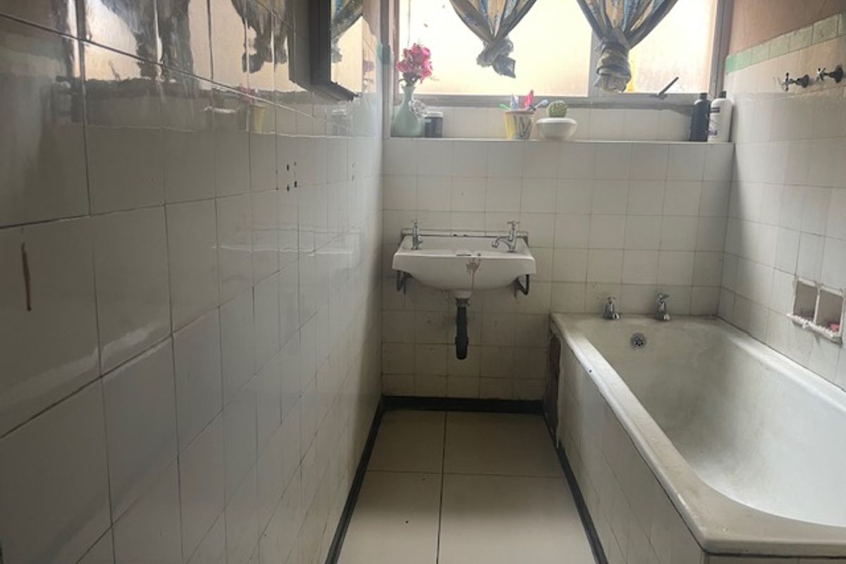 A dated bathroom featuring a wall-mounted basin, built-in bathtub, and full-height ceramic wall tiling, requiring modernization and cosmetic updates.