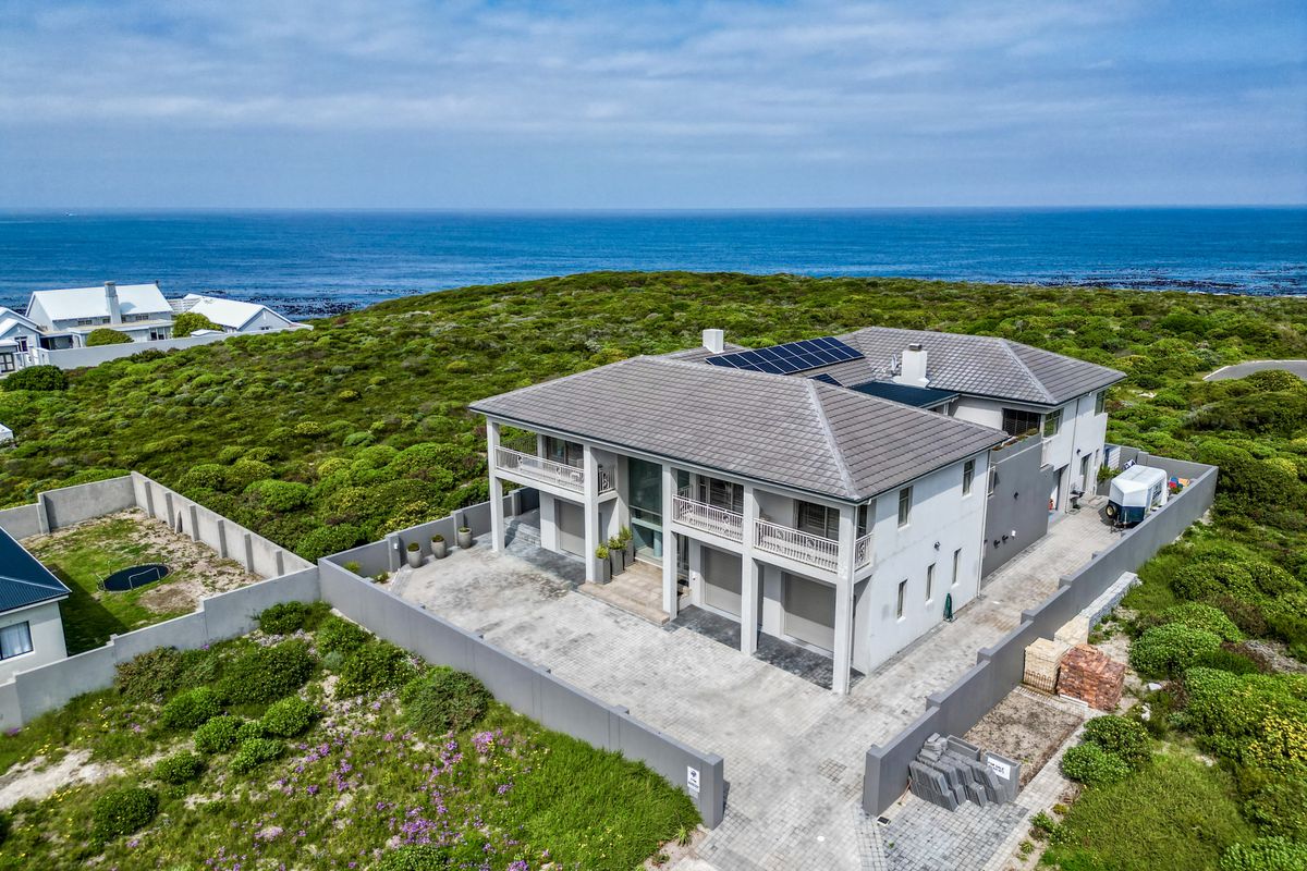 Aerial view of a modern two-story coastal residence featuring a tiled roof with solar panels, paved driveway, and multiple balconies overlooking the ocean.