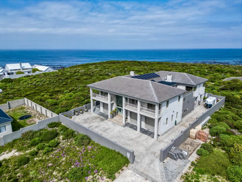 Aerial view of a modern two-story coastal residence featuring a tiled roof with solar panels, paved driveway, and multiple balconies overlooking the ocean.