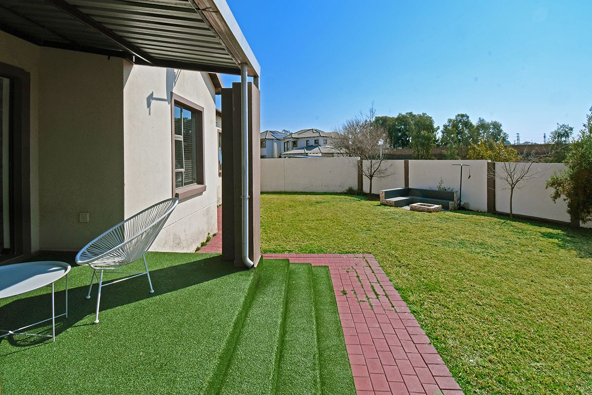 Rear exterior view featuring a covered patio with artificial grass, brick paving, a landscaped lawn, and a perimeter wall with built-in seating.