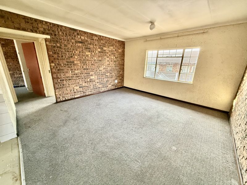 An empty living room featuring an exposed brick feature wall, carpeted flooring, and a large steel-framed window, requiring cosmetic renovation and updates.
