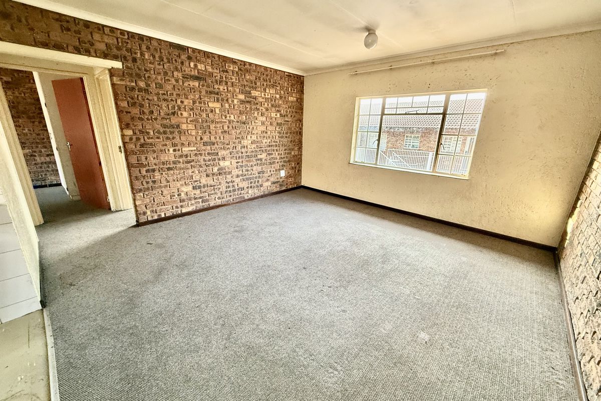 An empty living room featuring an exposed brick feature wall, carpeted flooring, and a large steel-framed window, requiring cosmetic renovation and updates.