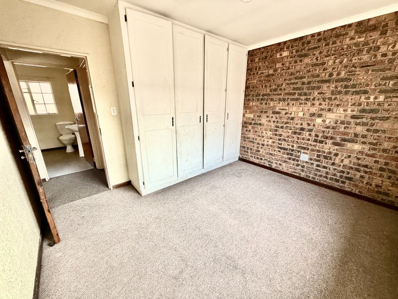 A bedroom featuring built-in cupboards and an exposed brick wall, with an adjacent bathroom visible through the doorway.