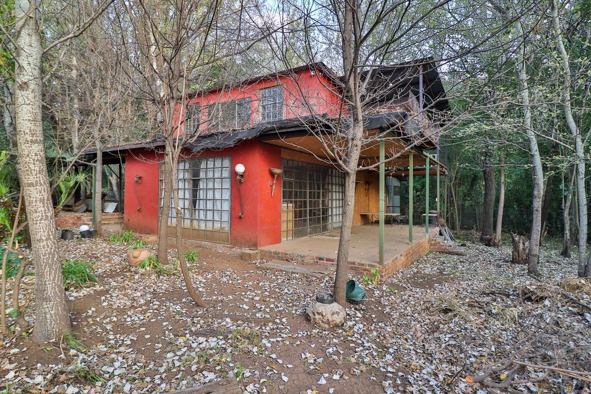 A two-story red brick dwelling featuring a covered patio and upper balcony, situated in a wooded area with visible signs of exterior wear.