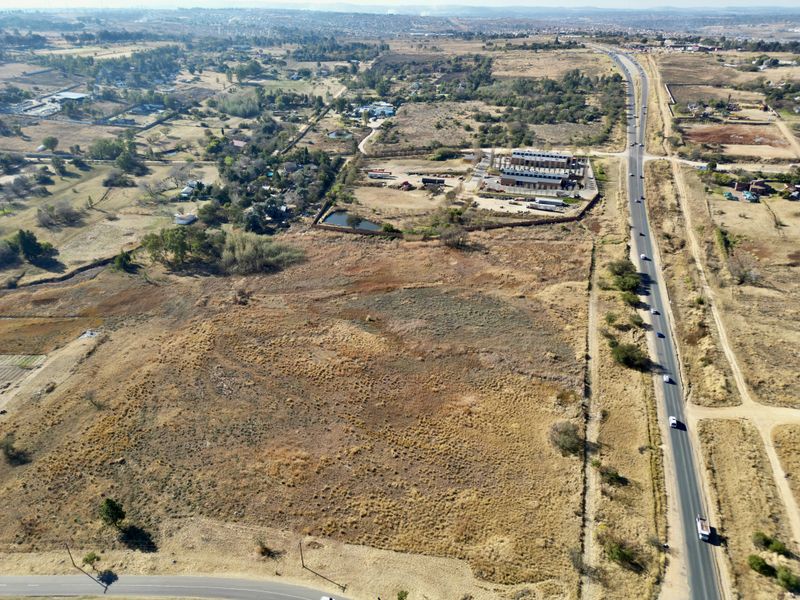 An aerial view of a large, undeveloped vacant residential plot situated adjacent to a main road with surrounding rural and semi-industrial landscape.