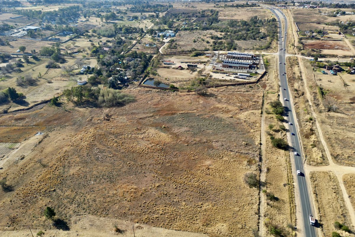 An aerial view of a large, undeveloped vacant residential plot situated adjacent to a main road with surrounding rural and semi-industrial landscape.