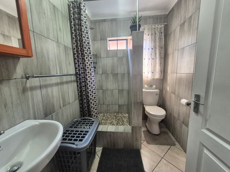 A compact bathroom featuring floor-to-ceiling grey wood-look wall tiles, a walk-in shower with pebble flooring, a pedestal basin, and a standard toilet.