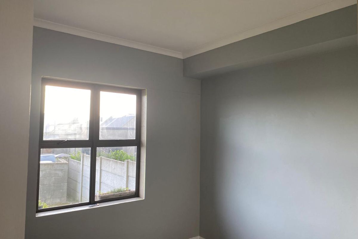 A bright, empty bedroom featuring neutral grey walls, coving, skirting boards, and an aluminium-framed window overlooking the exterior.