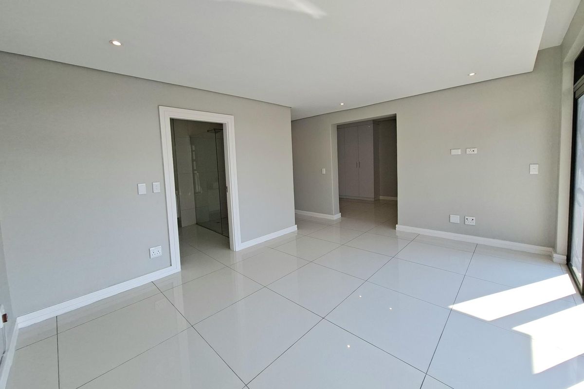 A bright, modern, and empty bedroom featuring neutral walls, large porcelain floor tiles, recessed lighting, and an en-suite bathroom access.