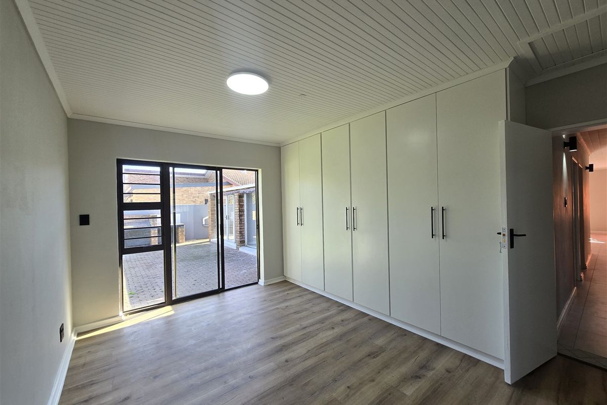 A bright, empty bedroom featuring modern laminate flooring, extensive built-in cupboards, and a sliding glass door providing access to the rear patio.