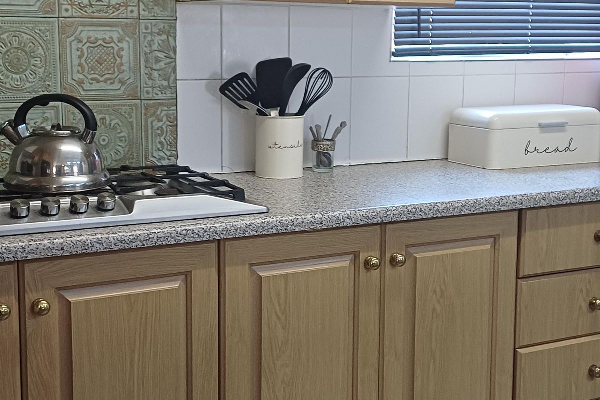 A functional kitchen featuring light wood-tone cabinetry, a gas cooktop with decorative tiled splashback, stone-look laminate countertops, and durable ceramic floor tiles.