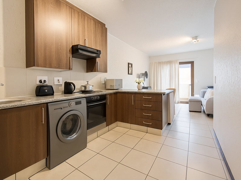 Open-plan kitchen featuring wood-finish cabinetry, granite countertops, built-in oven, and integrated laundry space, flowing into a bright living area with balcony access.