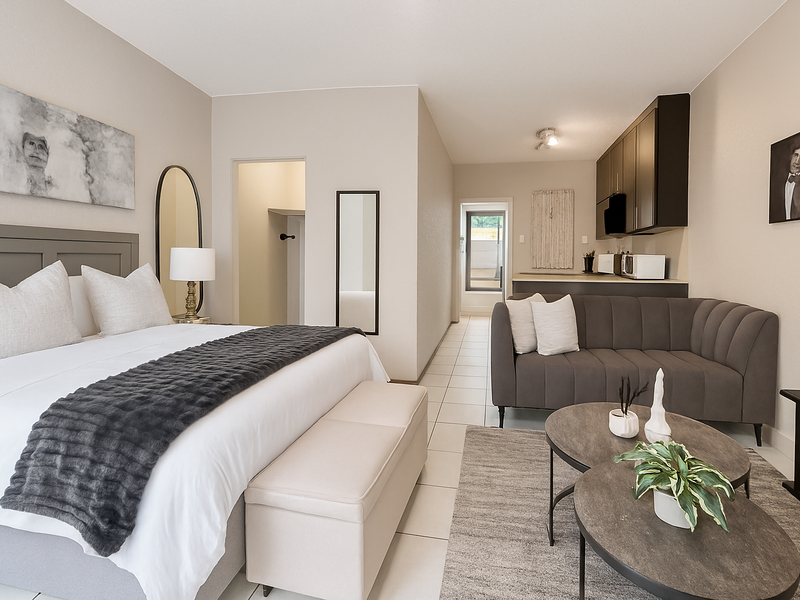 A studio apartment featuring a combined bedroom and living area with tiled flooring, neutral walls, and a compact kitchenette with dark cabinetry.