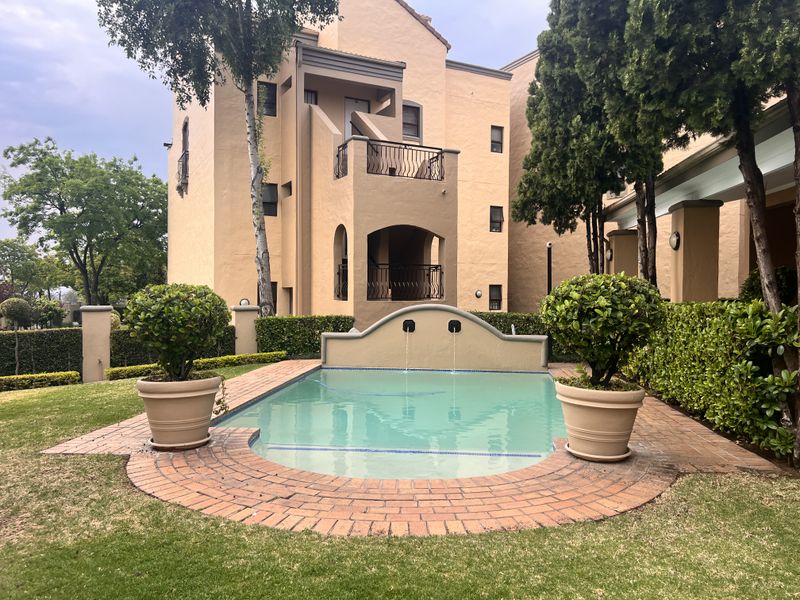 A well-maintained communal swimming pool area featuring a water fountain feature, brick paving, and landscaped garden surrounds within an apartment complex.