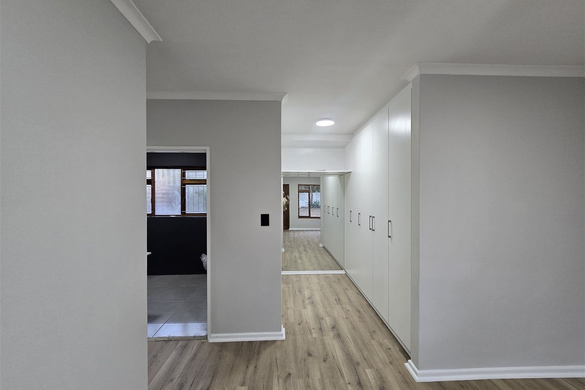 A bright, modern bedroom featuring extensive built-in cupboards with mirrored doors, sleek laminate flooring, and clean neutral finishes throughout.