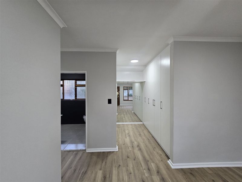 A bright, modern bedroom featuring extensive built-in cupboards with mirrored doors, sleek laminate flooring, and clean neutral finishes throughout.