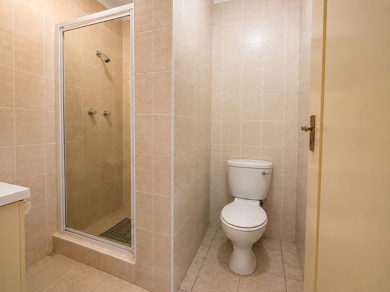 A functional bathroom featuring a tiled shower enclosure, a standard toilet, and a vanity cabinet with neutral wall and floor tiling.