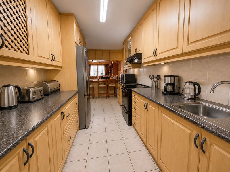 A functional galley-style kitchen featuring light wood cabinetry, dark laminate countertops, tiled flooring, and integrated appliances, leading into an open-plan living area.