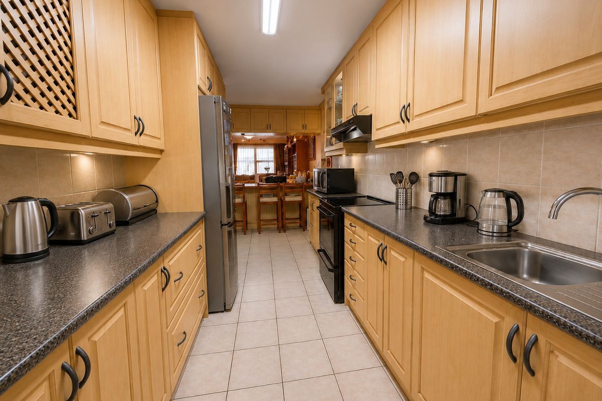 A functional galley-style kitchen featuring light wood cabinetry, dark laminate countertops, tiled flooring, and integrated appliances, leading into an open-plan living area.