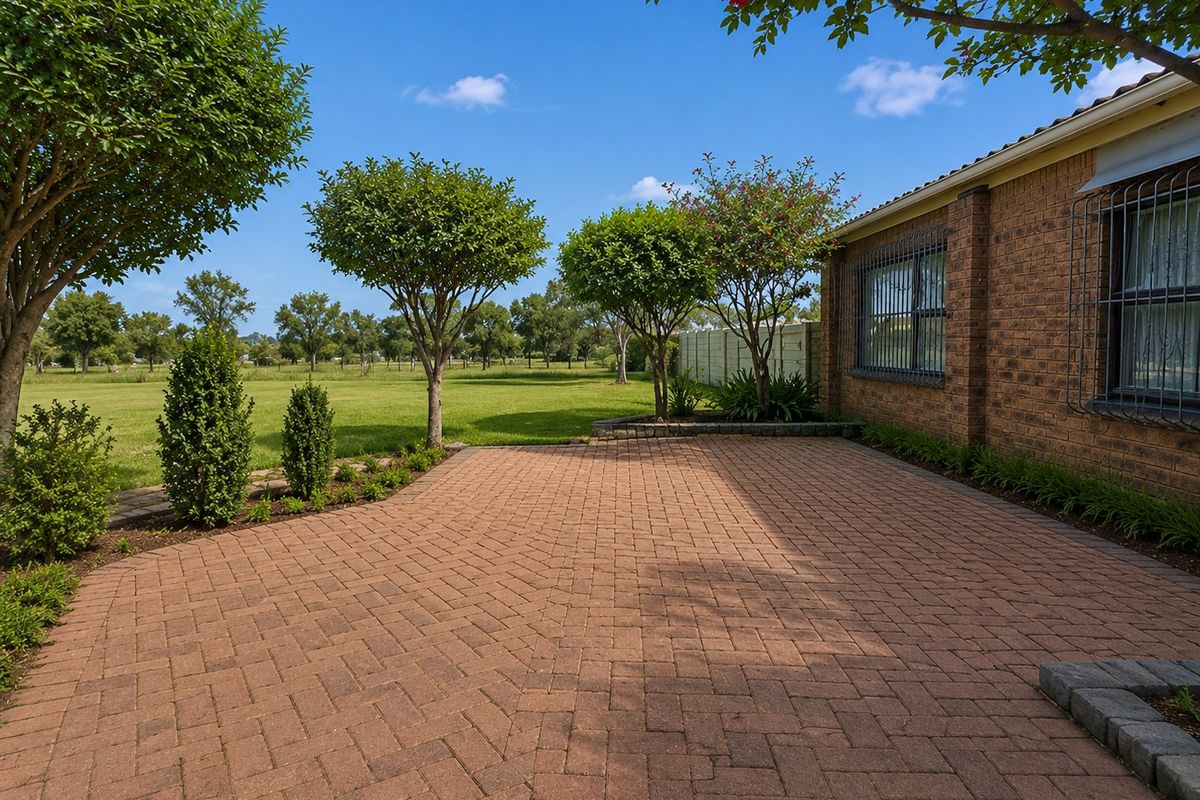 A well-maintained rear patio featuring brick paving, security bars on windows, and a landscaped garden overlooking an open green space.