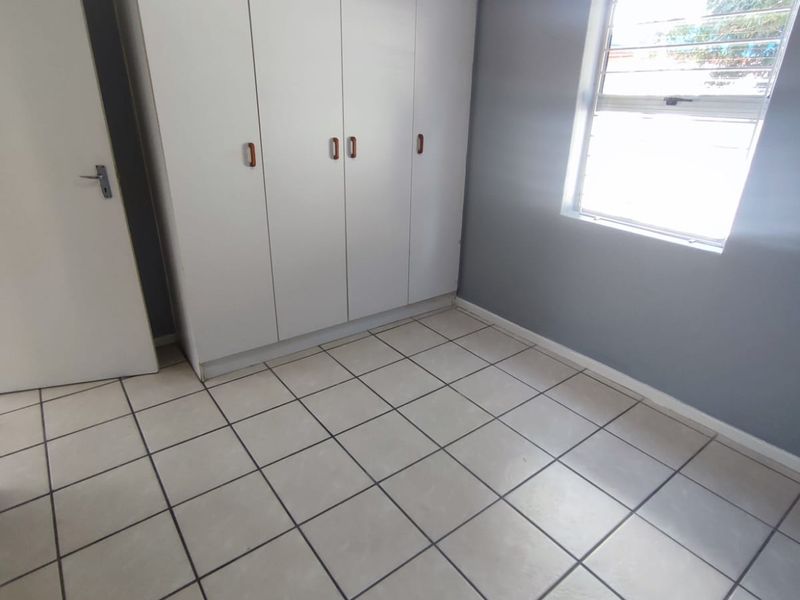 A bright, empty bedroom featuring neutral tiled flooring, built-in white cupboards, and a louver window.