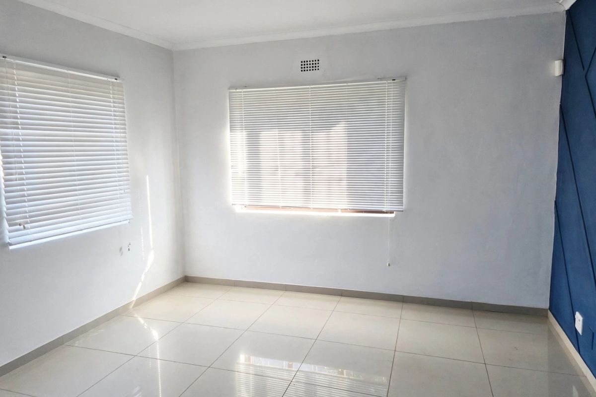 A bright, empty bedroom featuring neutral tiled flooring, white walls, venetian blinds, and a blue geometric accent wall.