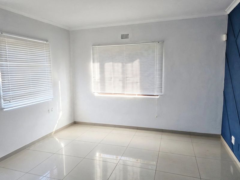 A bright, empty bedroom featuring neutral tiled flooring, white walls, venetian blinds, and a blue geometric accent wall.