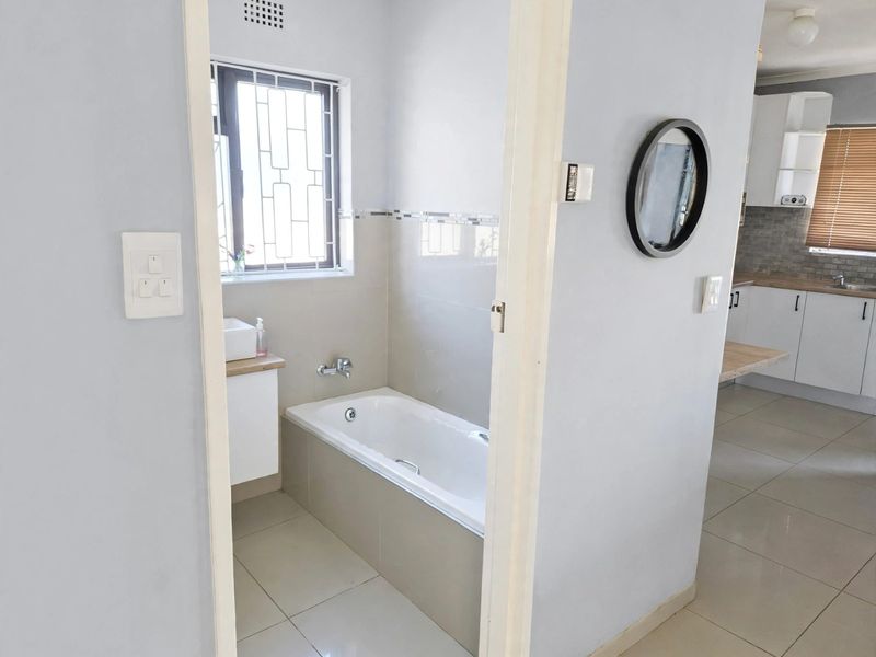 A clean, functional bathroom featuring a built-in bathtub, vanity cabinet, and tiled flooring, with a view into the adjacent kitchen area.