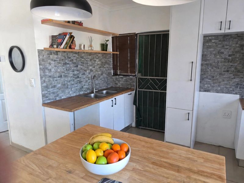 A functional kitchen featuring white cabinetry, wooden countertops, a stone-look tile backsplash, and floating shelves, with a security gate leading to the exterior.