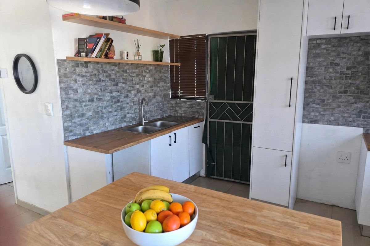 A functional kitchen featuring white cabinetry, wooden countertops, a stone-look tile backsplash, and floating shelves, with a security gate leading to the exterior.