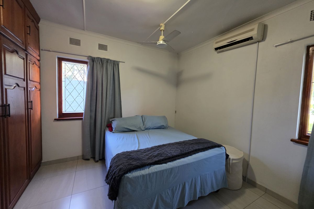 A functional bedroom featuring tiled flooring, a built-in wooden wardrobe, ceiling fan, and a wall-mounted split-system air conditioner.
