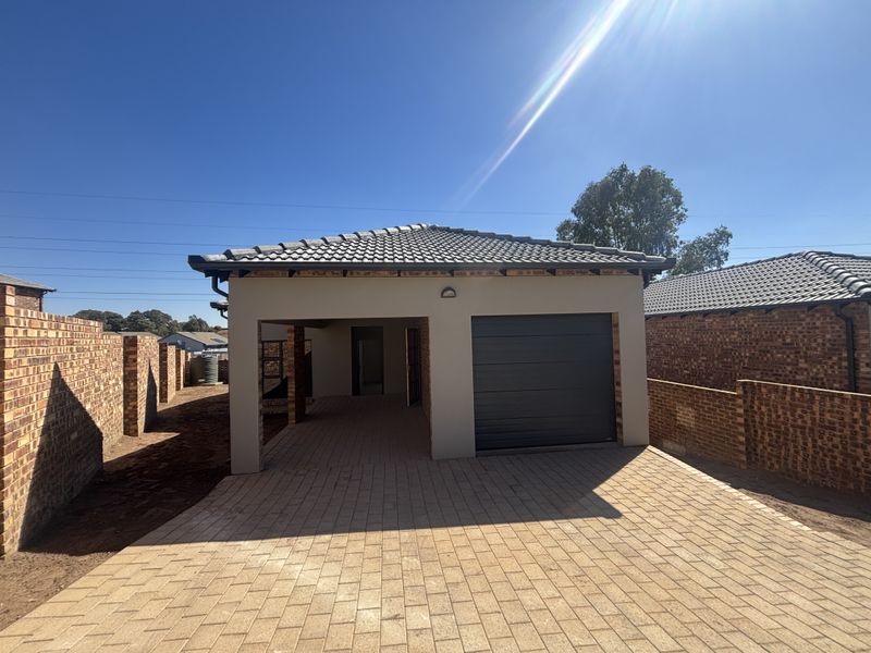 A modern residential exterior featuring a paved driveway, a single garage, and a covered carport area, enclosed by face-brick boundary walls.