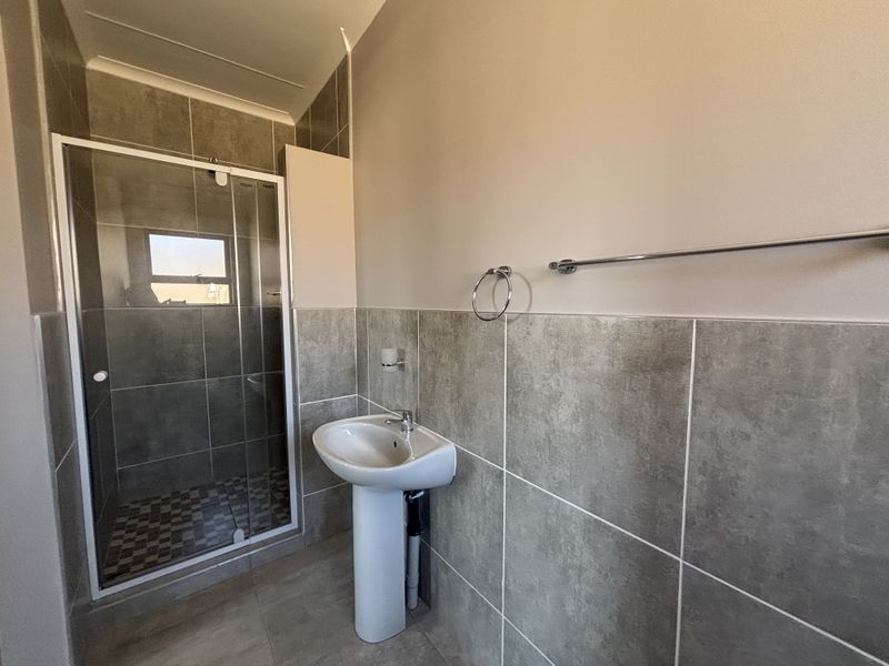 A clean, modern bathroom featuring a pedestal basin, a glass-enclosed shower, and neutral grey wall and floor tiling.