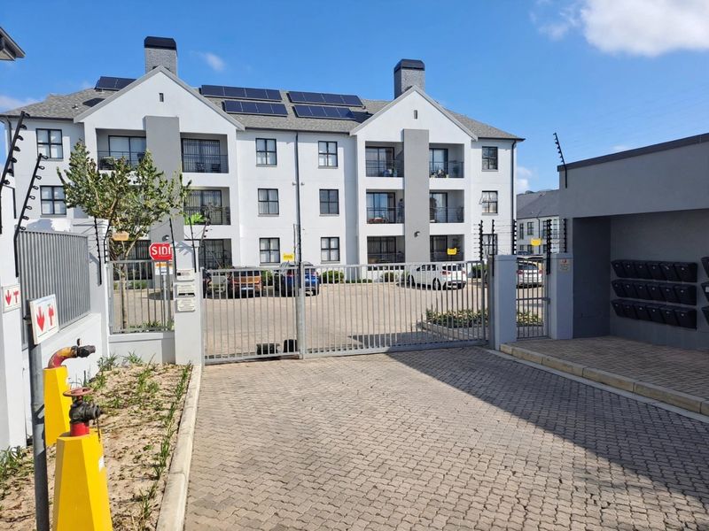 Front view of a modern apartment complex featuring a secure gated entrance, paved driveway, solar panels, and integrated post boxes.