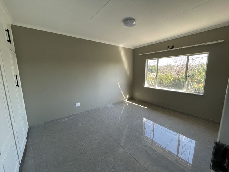 A bright, empty bedroom featuring modern grey tiled flooring, built-in cupboards, and a window with security bars overlooking the exterior.
