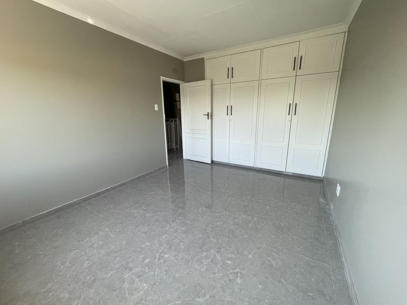 A clean, empty bedroom featuring modern grey tiled flooring, neutral wall paint, and extensive white built-in cabinetry with overhead storage.