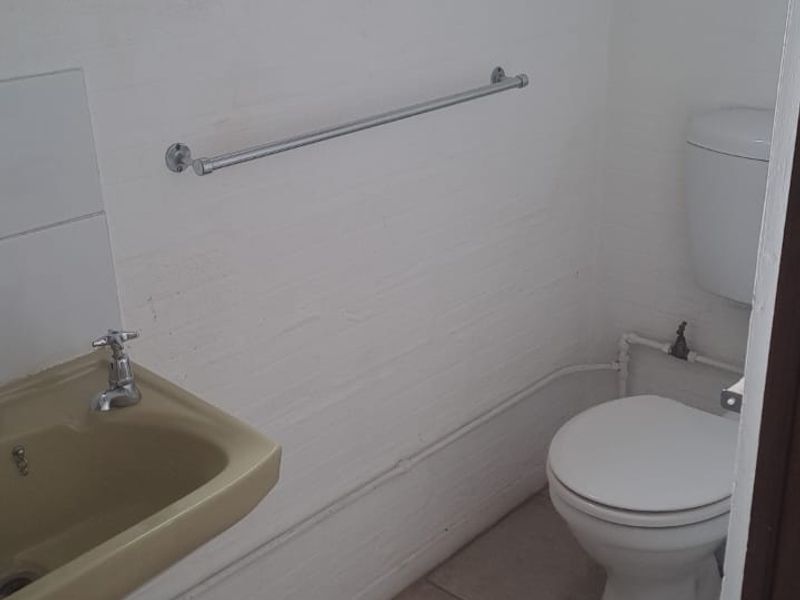 A compact bathroom featuring a white toilet, a vintage-style washbasin, and tiled flooring, with exposed plumbing and a functional top-hung window.