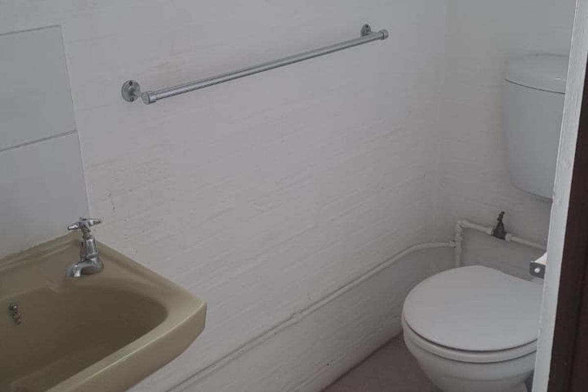 A compact bathroom featuring a white toilet, a vintage-style washbasin, and tiled flooring, with exposed plumbing and a functional top-hung window.