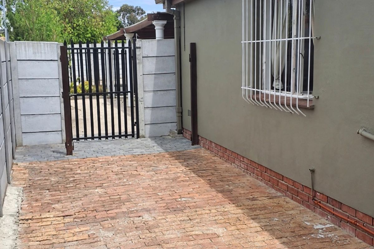 A rear exterior area featuring brick paving, a secure metal gate, and a plastered wall with security bars on the window.