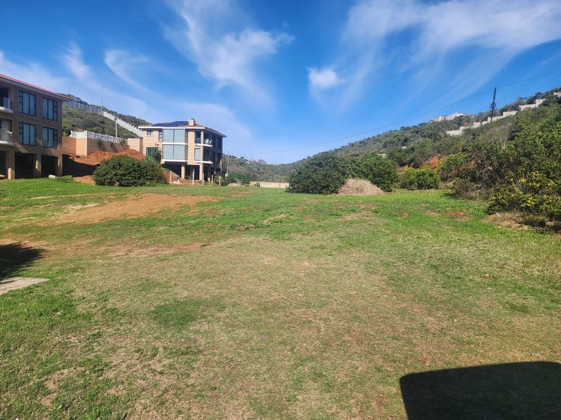 A view of a vacant residential plot featuring sloped grassy terrain with surrounding hillside landscape and neighboring residential properties.