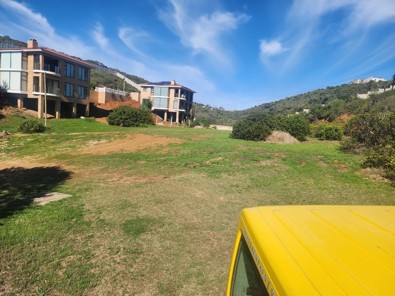 A view of a vacant residential plot featuring sloped grassy terrain with neighboring multi-story residential properties in the background.