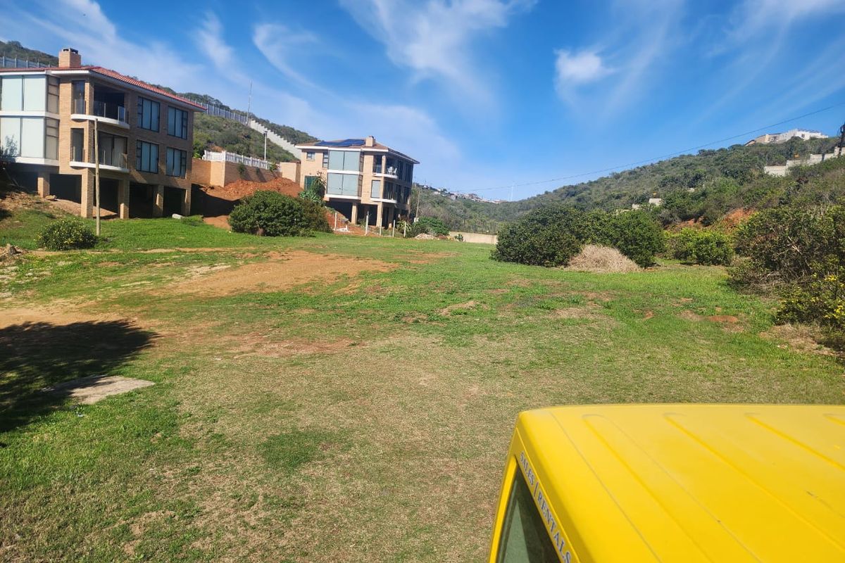 A view of a vacant residential plot featuring sloped grassy terrain with neighboring multi-story residential properties in the background.