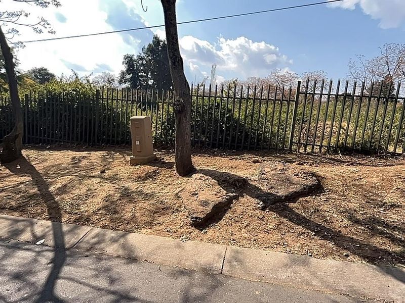 A view of the vacant residential land plot featuring perimeter metal fencing, mature trees, and an electrical utility box.