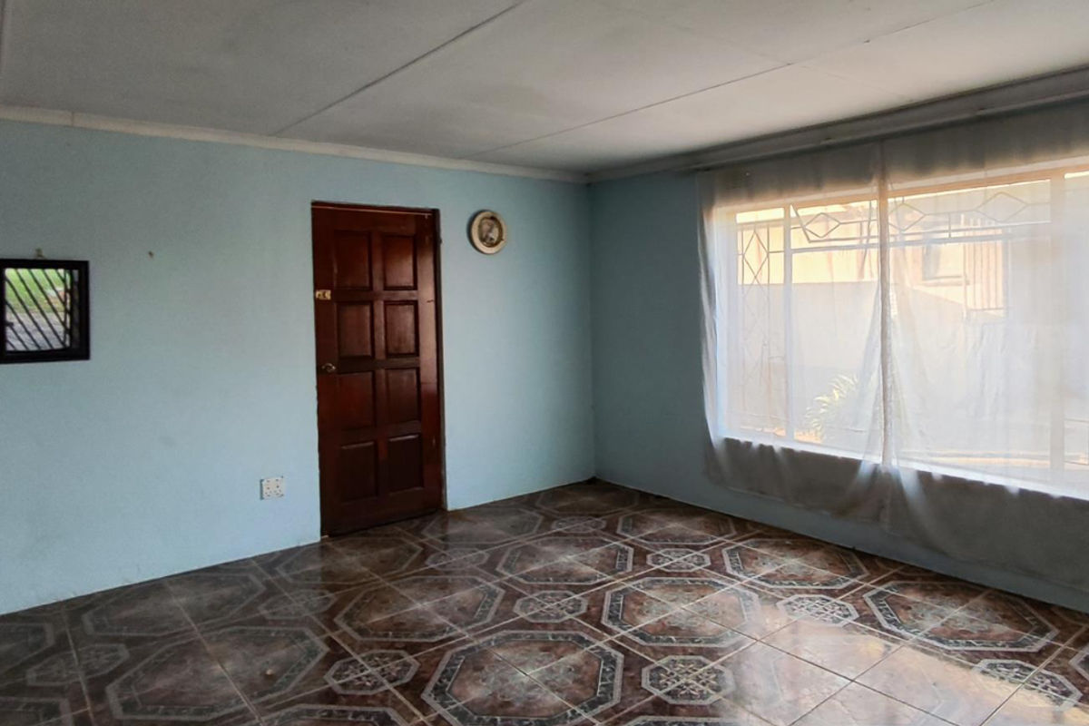 An empty living room featuring patterned floor tiles, a wooden door, and a large window with security bars.
